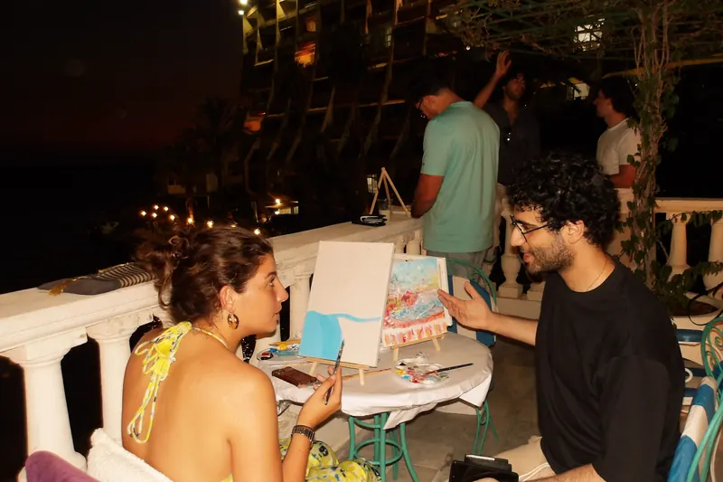 Two guests at their easels on a lamplit terrace, the bay dark behind them