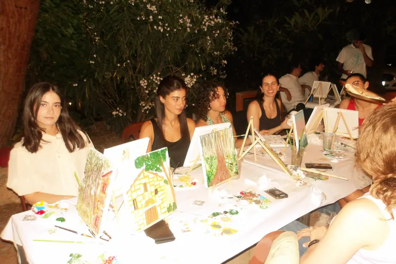 A long table of guests painting side by side under the trees after dark