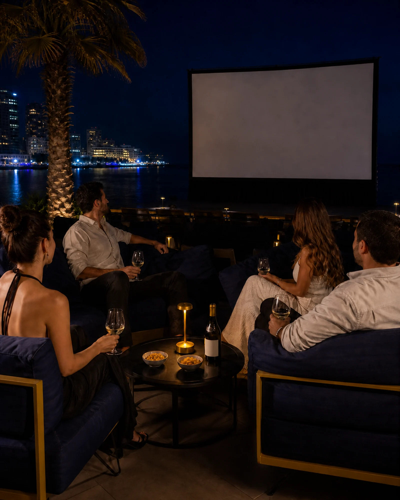 Four adults in navy lounge armchairs at Boha, around a low table with nuts and one shared wine bottle, facing the outdoor cinema screen over the water.
